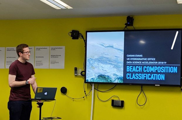 The author stood in front of monitor with slides displaying "Ciaran Evans, UK Hydrographic Office, Data Science Accelerator, Beach Composition Classification"